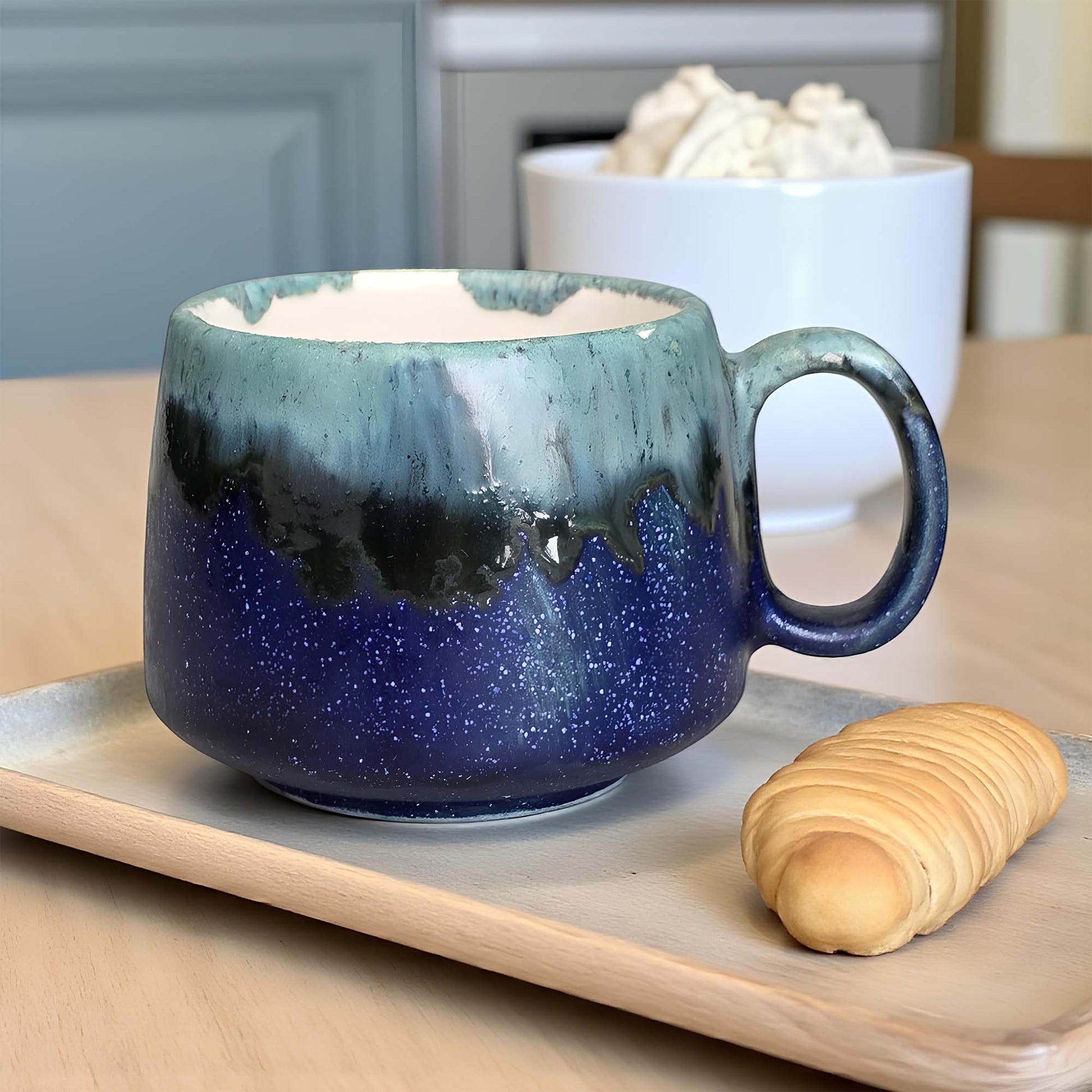 Blue ceramic mug with a handle on a wooden tray, with a bowl of whipped cream and a croissant in the background.