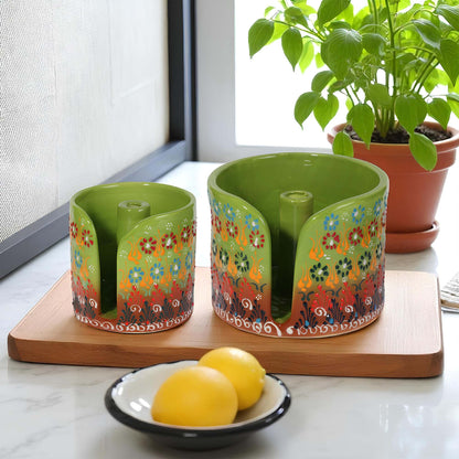 Two colorful ceramic paper towel holders with floral designs on a wooden board, with a bowl of lemons and a potted plant in the background.