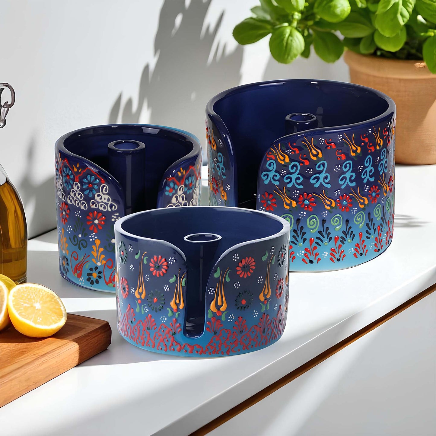 Three blue ceramic paper towel holders with colorful patterns on a white surface with a plant and lemon in the background.