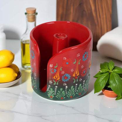 Red ceramic paper towel holder with floral patterns on a kitchen counter with lemons and a bottle in the background.