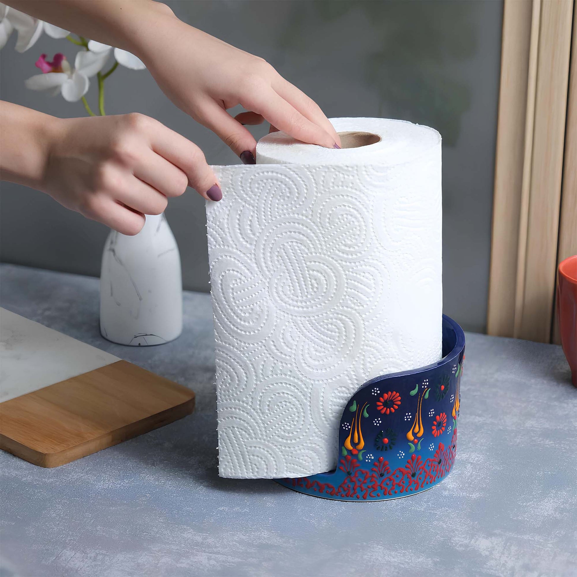 Person reaching for a roll of paper towels in a decorative paper towel holder on a kitchen counter.