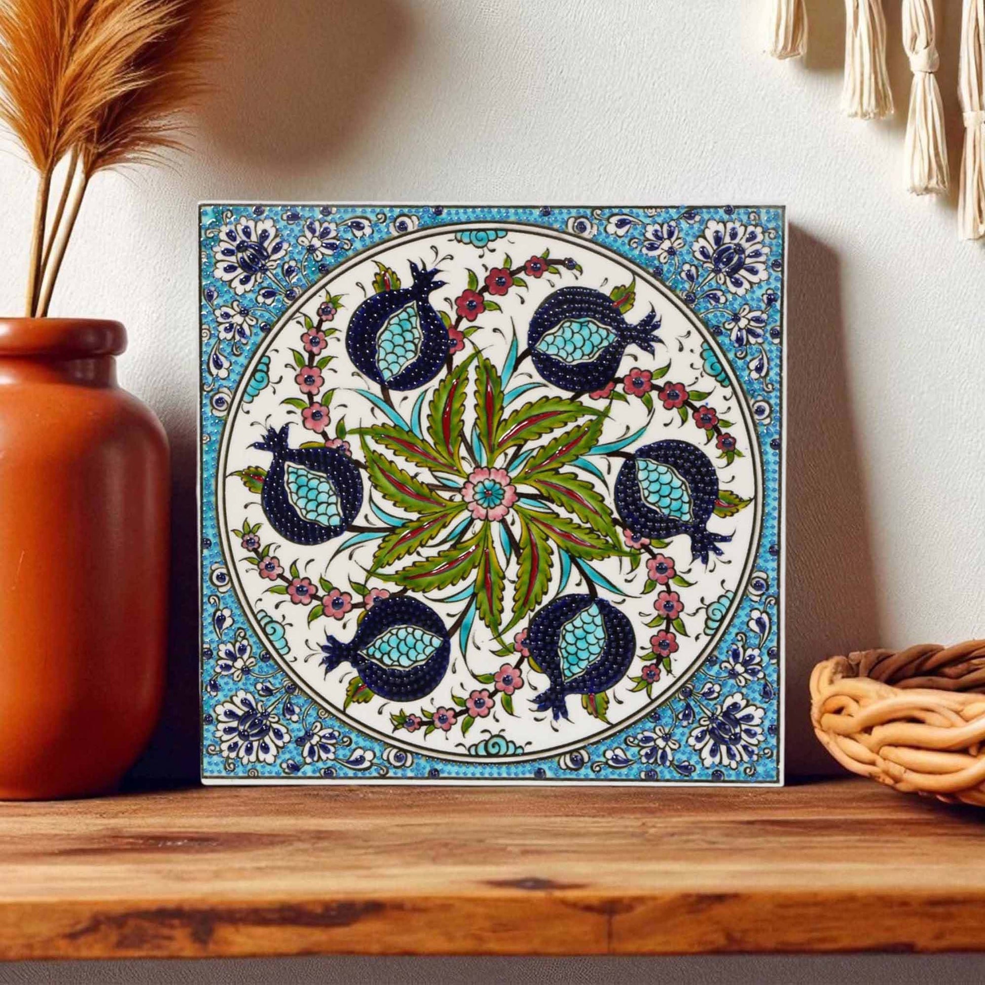 Decorative ceramic tile with floral and pomegranate design on a wooden shelf.