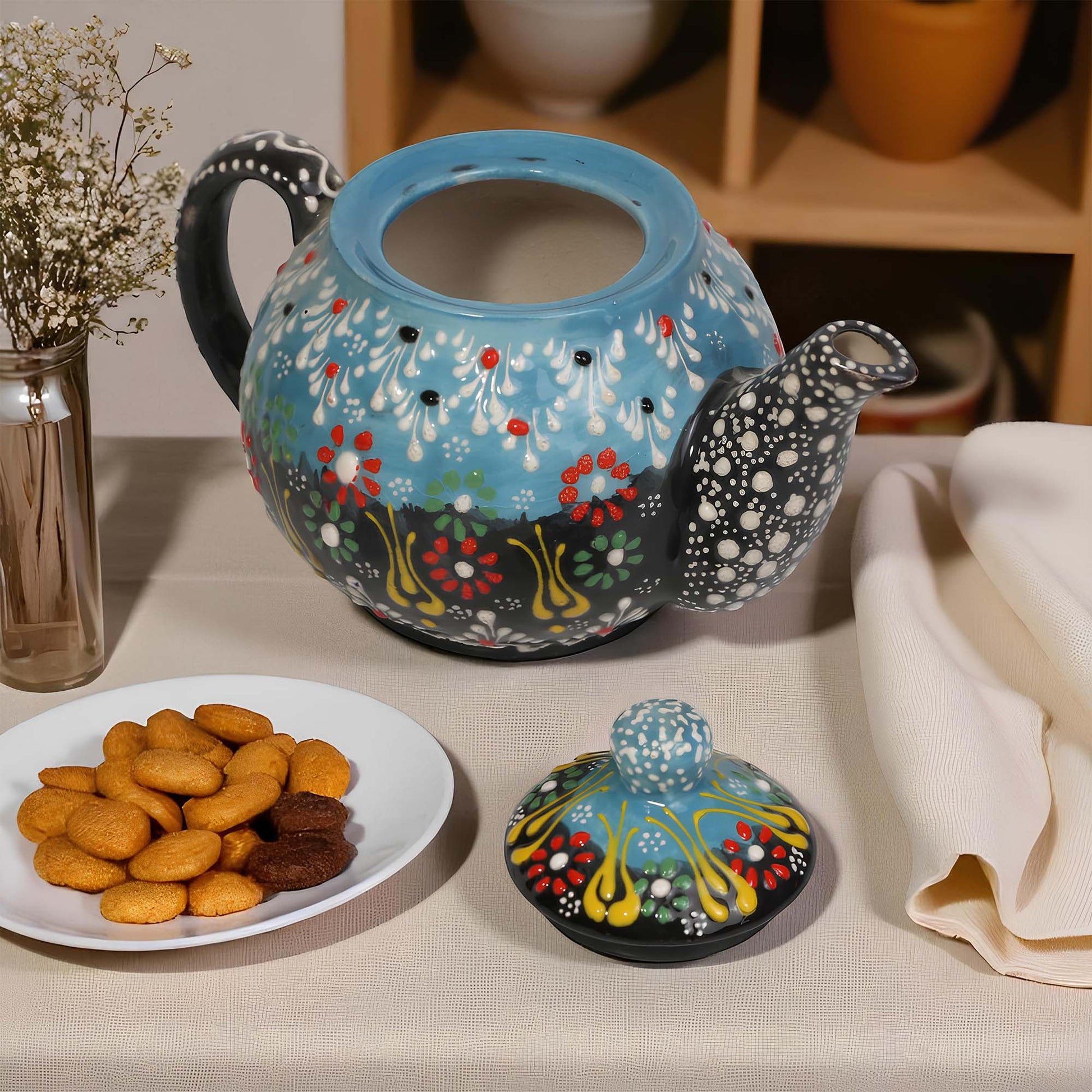 Decorative teapot with colorful floral pattern on a table with cookies and a vase.