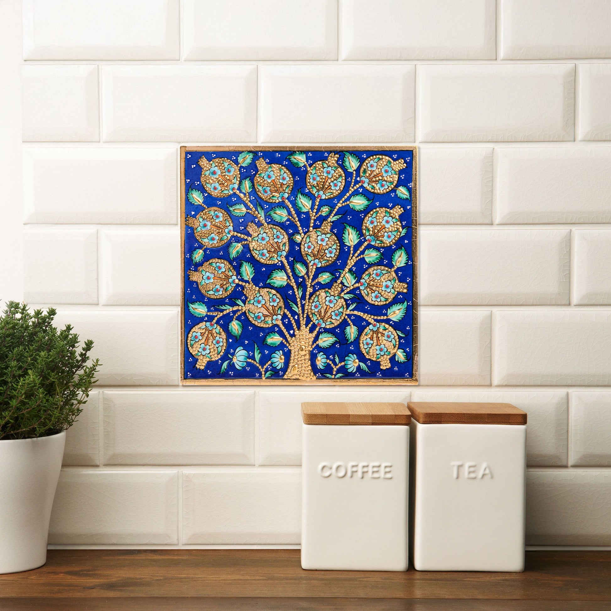 Decorative ceramic tile with floral tree design on a blue background, beside white kitchen canisters.