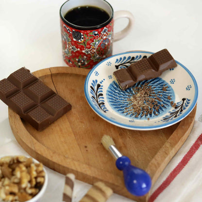 Chocolate bars on a wooden board with a mug of coffee and a decorative ceramic grater plate in the background.