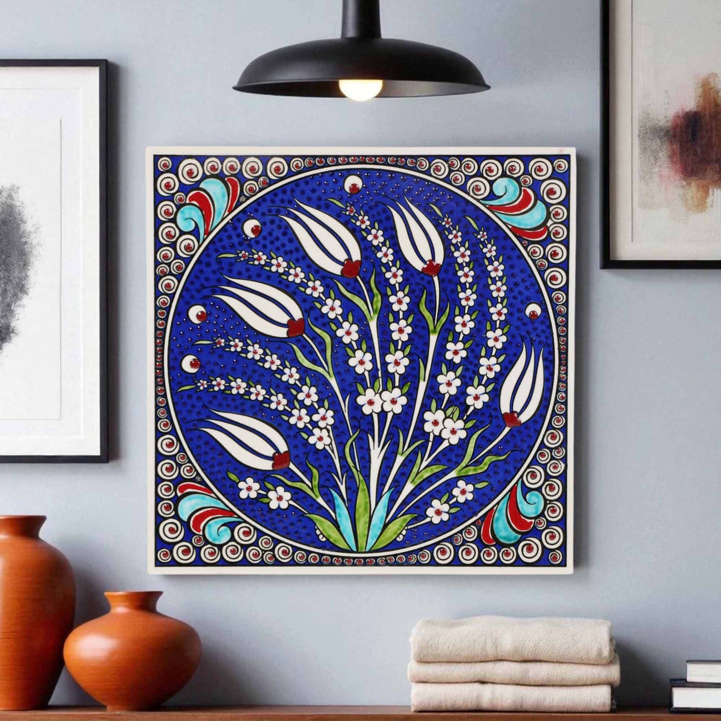 Decorative ceramic tile wall art with floral design on a blue background, framed against a gray wall with vases and books.