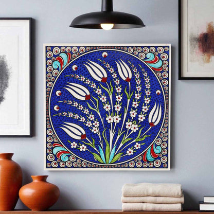 Decorative ceramic tile wall art with floral design on a blue background, framed against a gray wall with vases and books.