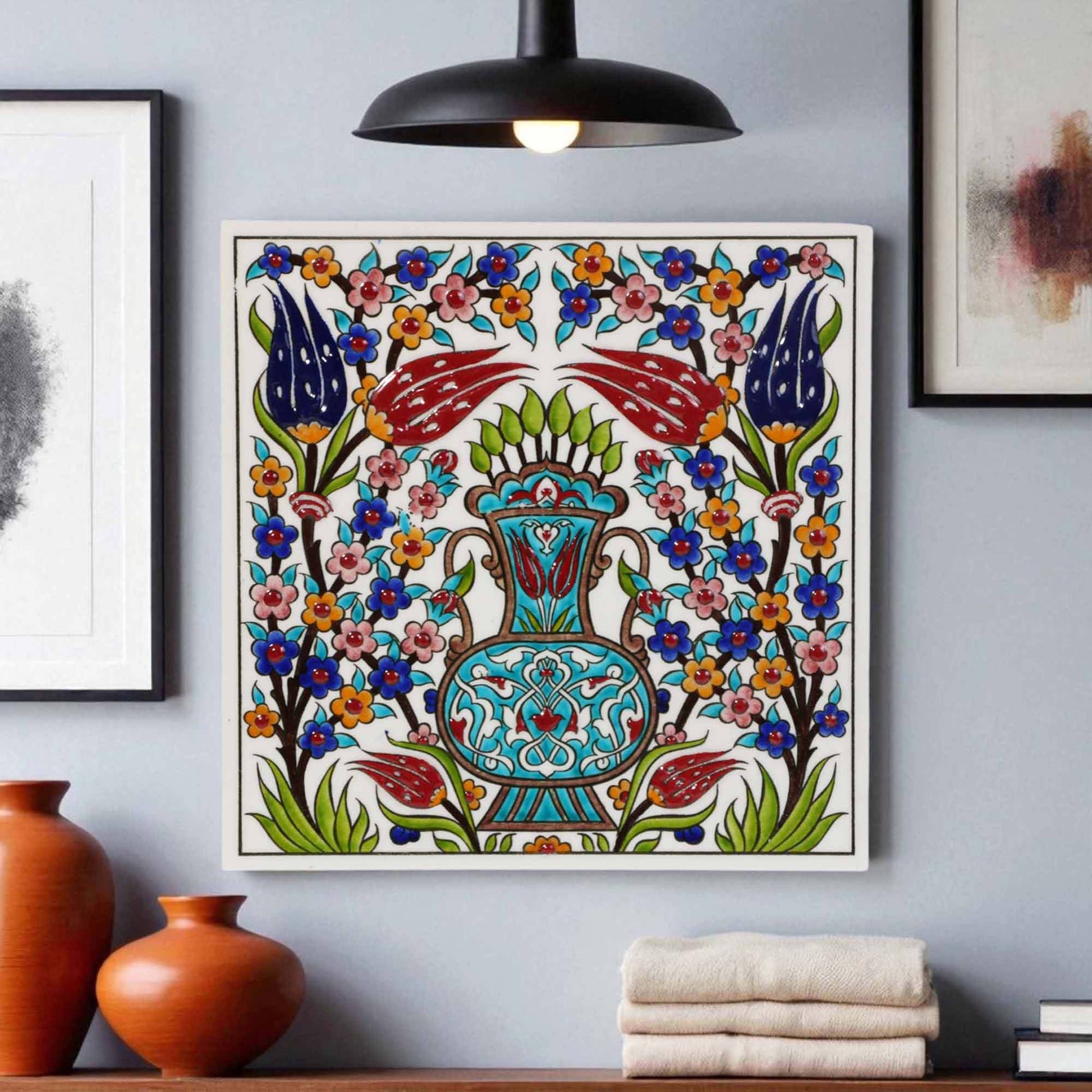 Decorative ceramic tile wall art with floral and chair design on a gray wall, with vases and books in the foreground.