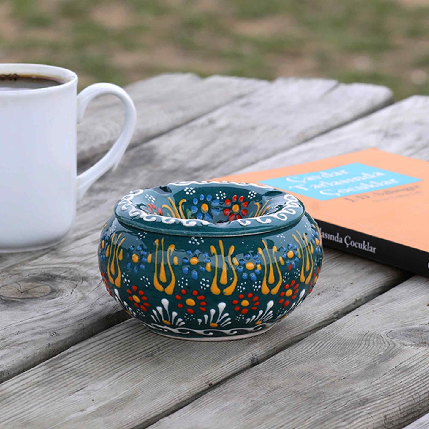 Decorative ceramic ashtray with floral patterns on a wooden table with a cup of coffee and book.