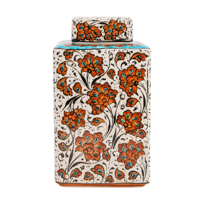 A square-shaped, hand-painted ceramic jar with a floral pattern in terracotta-orange blossoms with turquoise and black detailing on a white background, accompanied by its lid.