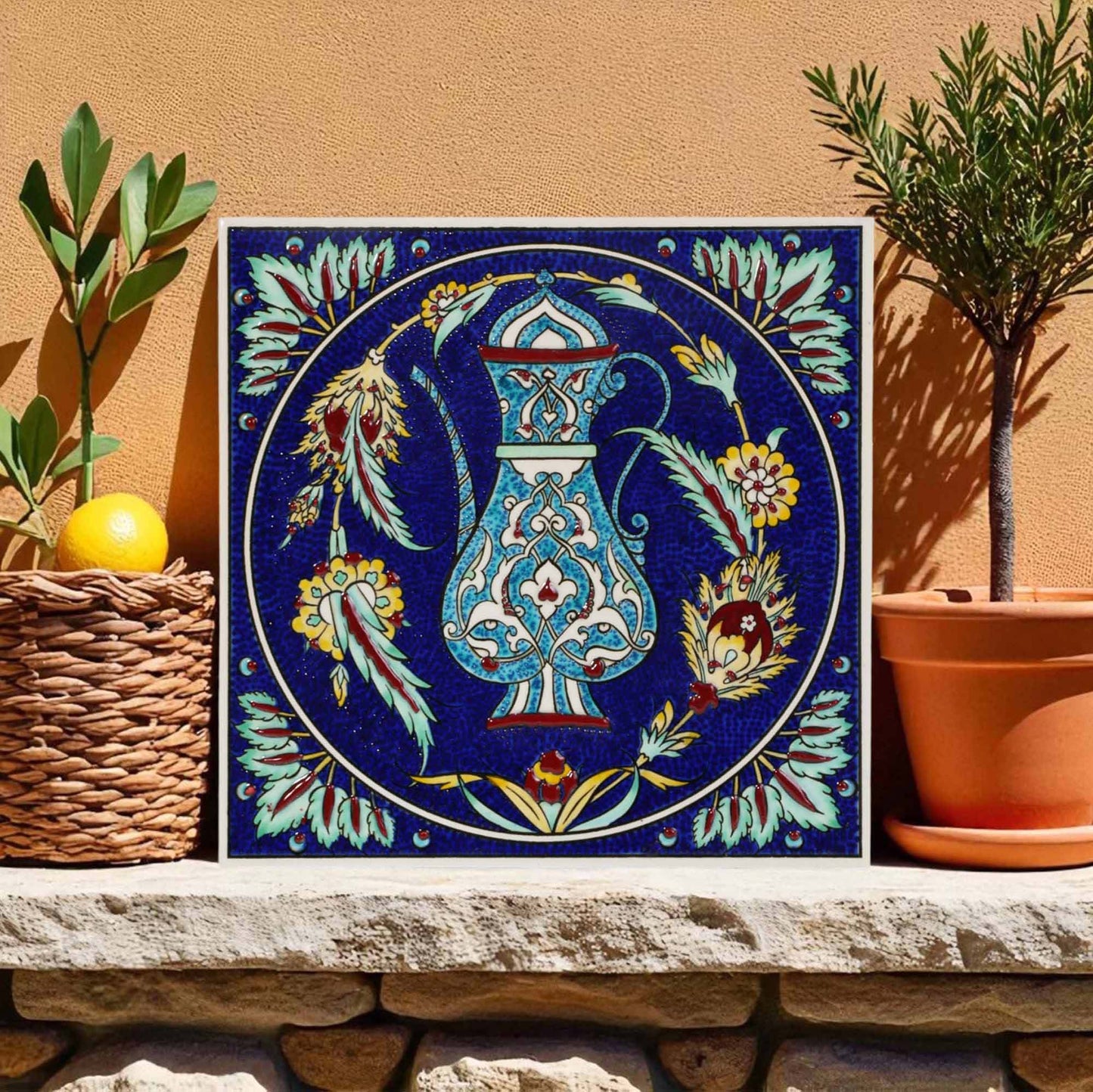 Decorative ceramic tile with floral and pitcher design on a stone ledge with plants.