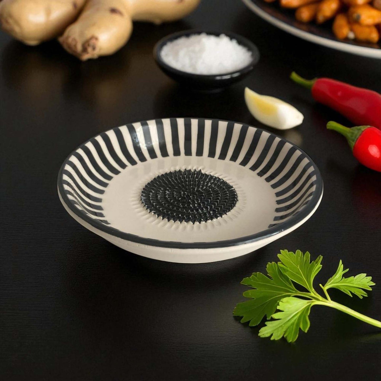 Black and white striped ceramic garlic grater bowl on a dark surface with ingredients around it.