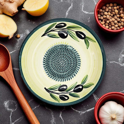 Decorative ceramic garlic grater bowl with olive design on a dark surface with kitchen items.