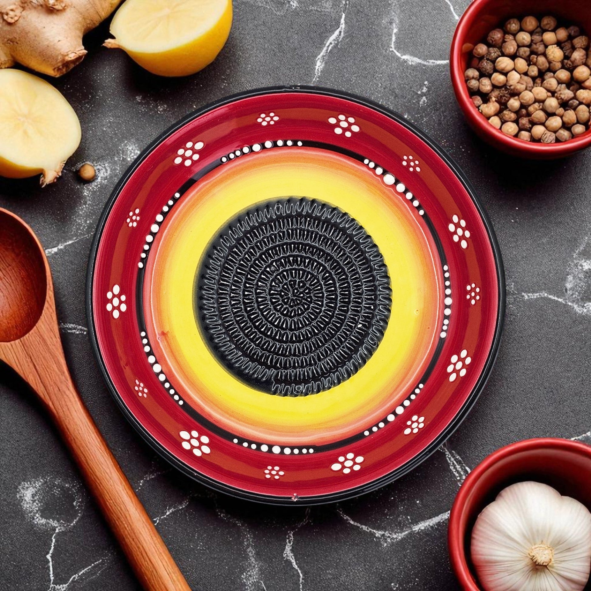 Decorative garlic grater bowl with a central black circular design on a red rim, surrounded by kitchen items on a dark surface.