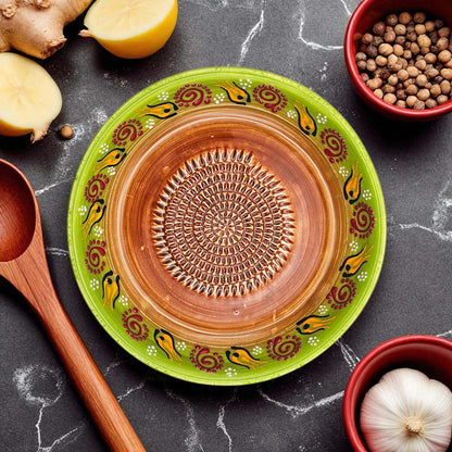 Ceramic garlic grater bowl with decorative green rim and brown center on a dark surface with kitchen items.