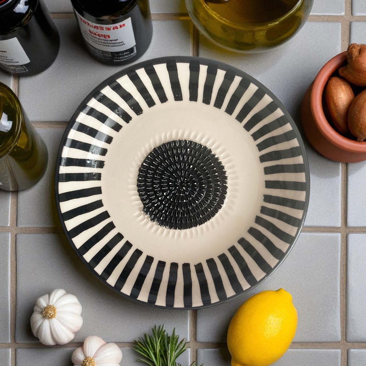 Decorative garlic grater bowl with black and white stripes on a tiled surface with lemons and bottles.