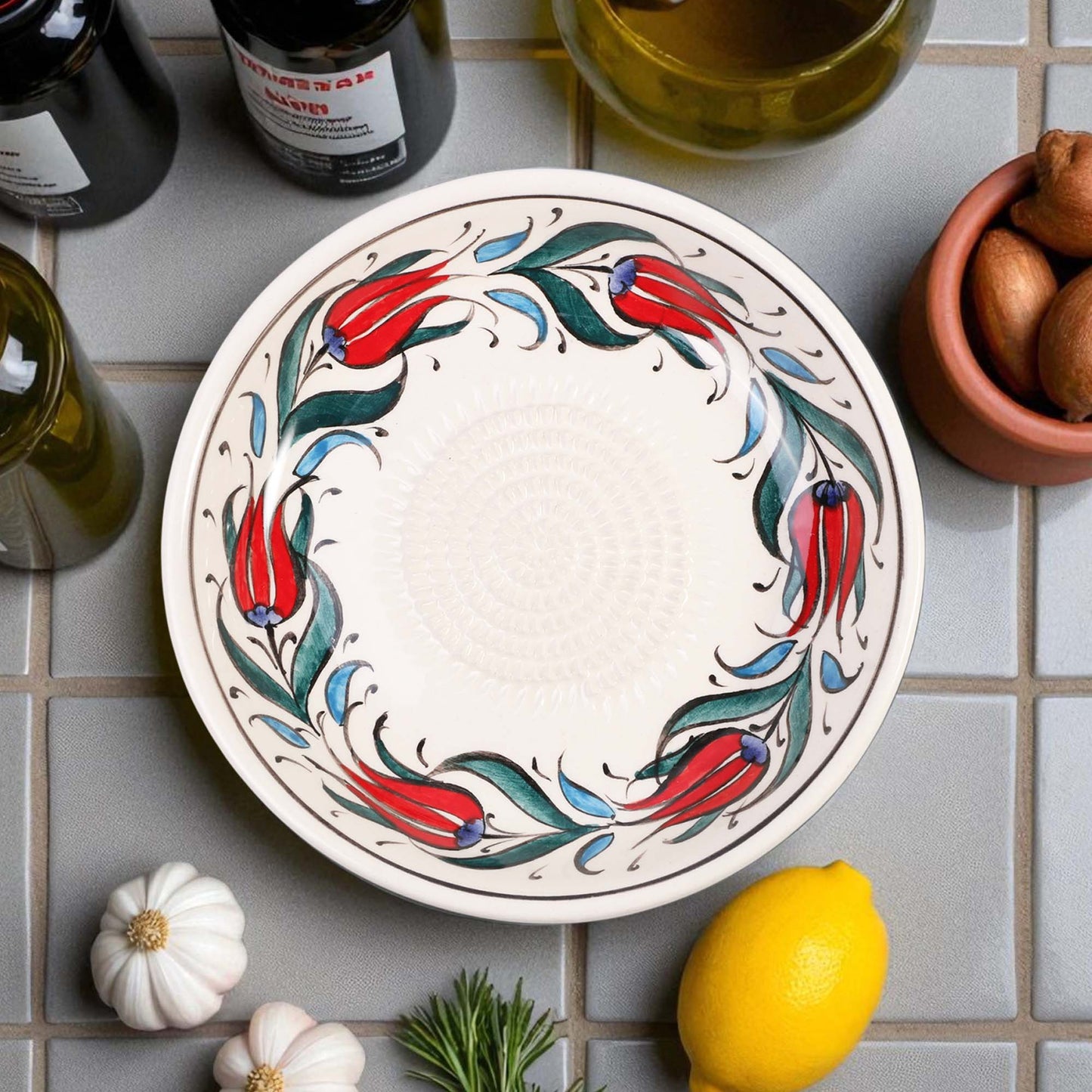 Decorative ceramic garlic grater bowl with floral design on a tiled surface with lemons and bottles.