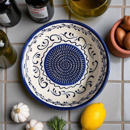 Decorative ceramic garlic grater bowl with blue center and white border on a tiled surface with lemons and garlic.