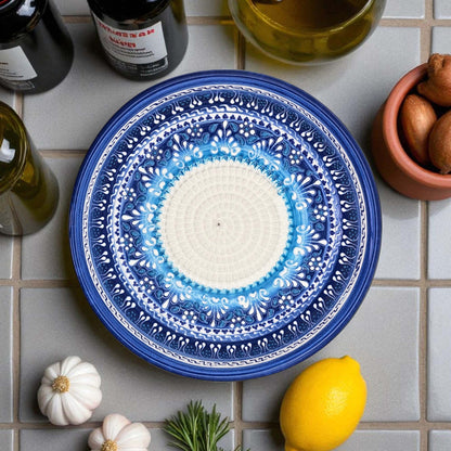 Blue decorative garlic grater bowl on a tiled surface with lemons, garlic, and bottles in the background