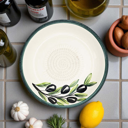 Decorative garlic grater bowl with olive design on a tiled surface with ingredients.
