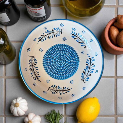 Decorative garlic grater bowl with blue and black patterns on a tiled surface with lemons and bottles.