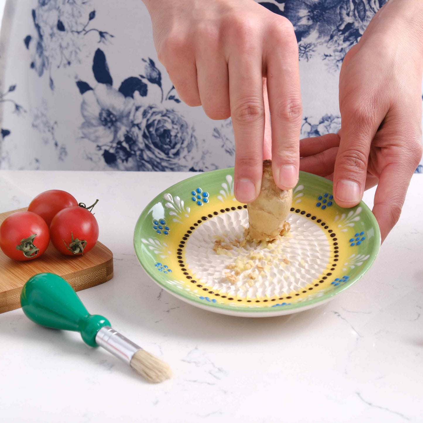 Person peeling a ginger root over a decorative ceramic garlic grater plate with tomatoes and a brush in the foreground.