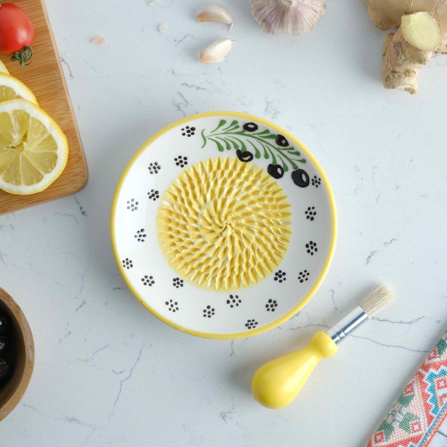 Decorative ceramic garlic grater plate with a yellow center on a white surface with kitchen items around