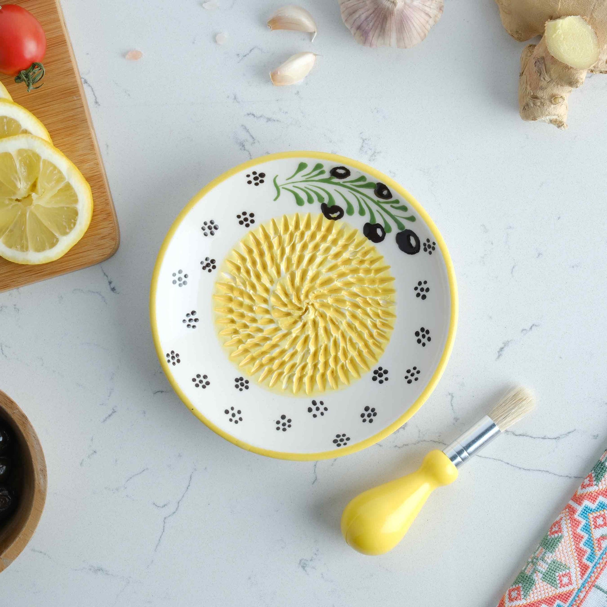 Decorative ceramic garlic grater plate with a yellow center on a white surface with kitchen items around
