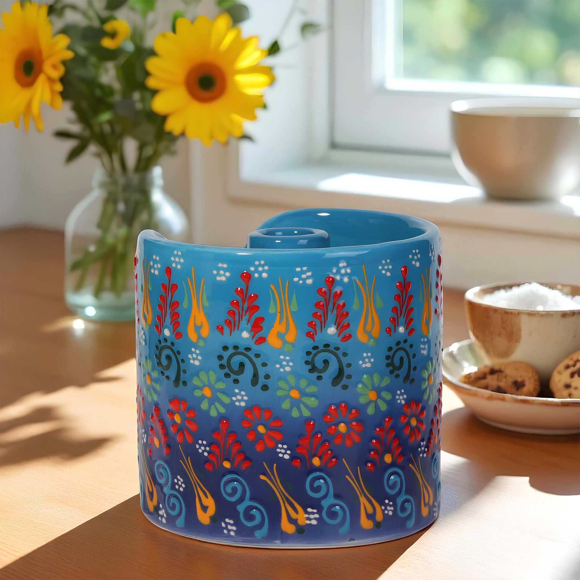 Blue ceramic paper towel holder with colorful floral pattern on a table with sunflowers and cookies.