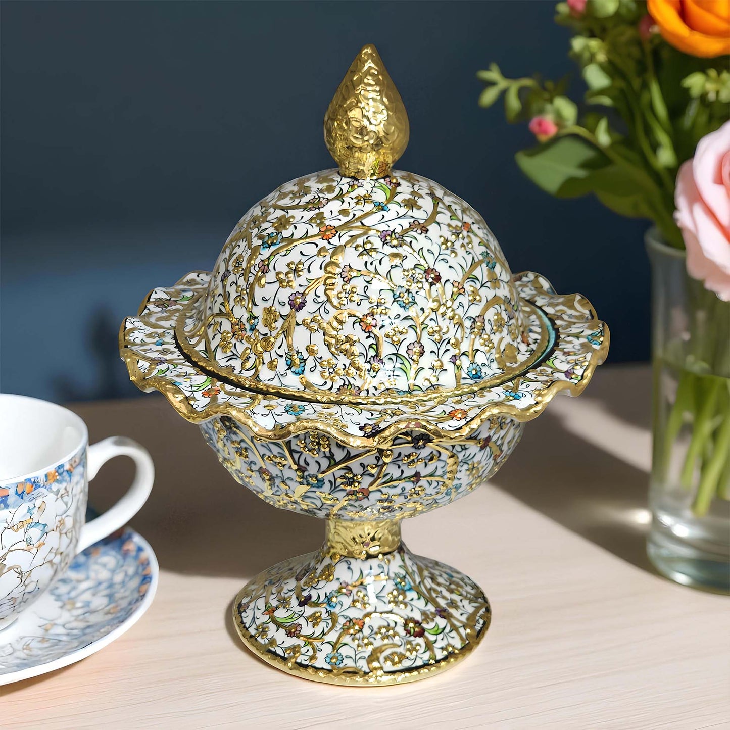 Decorative sugar bowl with intricate designs on a table with a cup and flowers in the background.