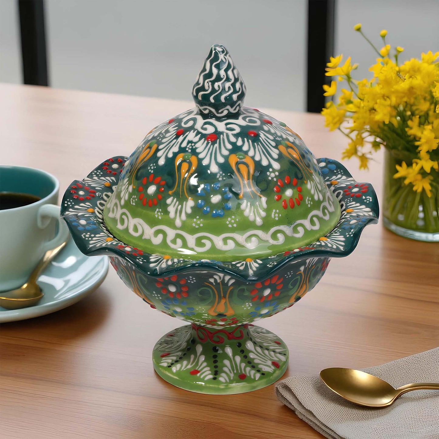 Decorative ceramic sugar bowl with a colorful lid on a table with a cup of coffee and flowers.