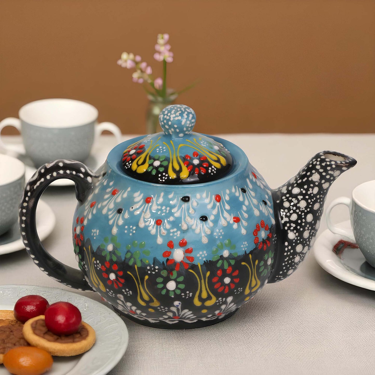 Decorative ceramic teapot with floral patterns on a table with cups and cookies.