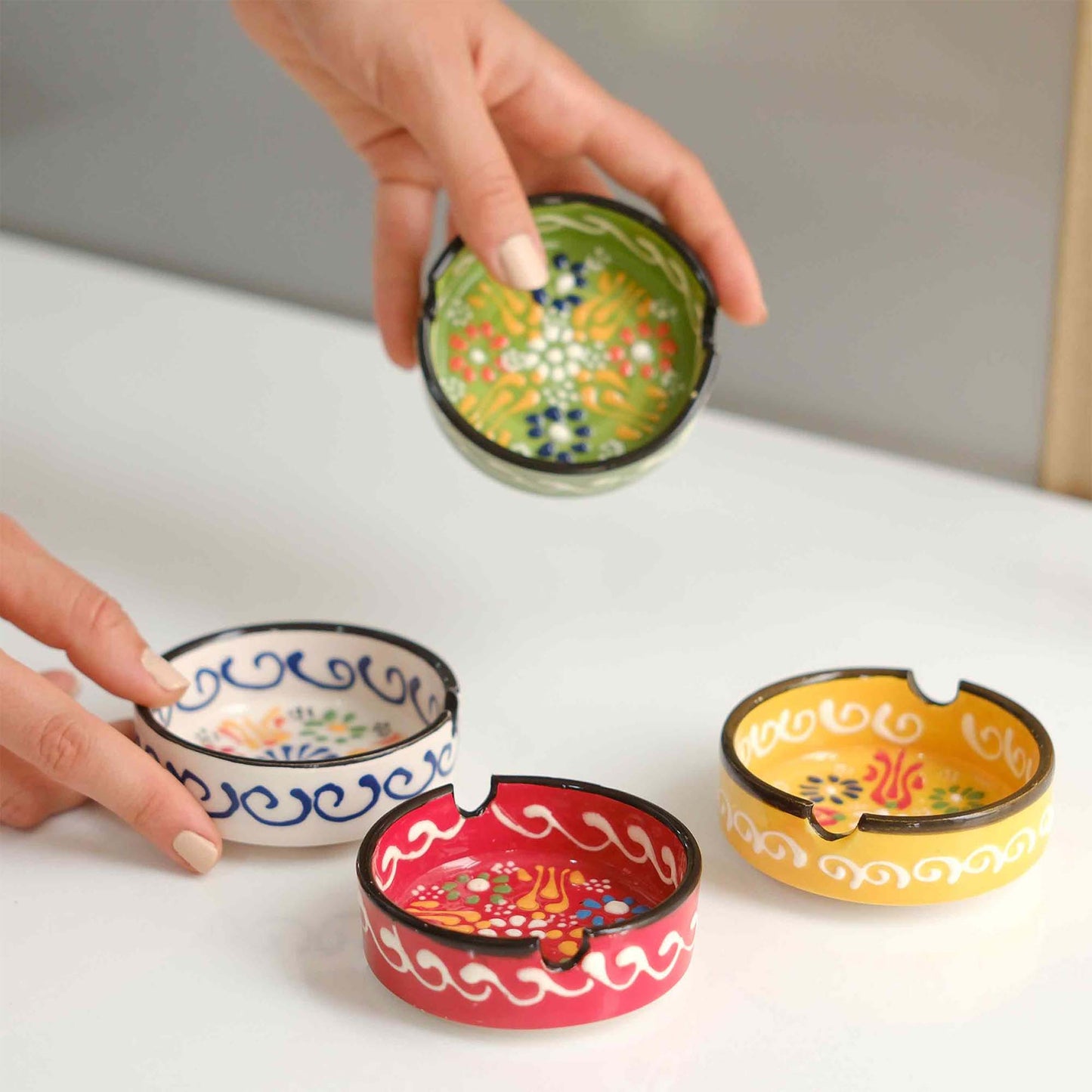 Set of colorful ceramic ashtrays on a white surface with a hand interacting with one of them.