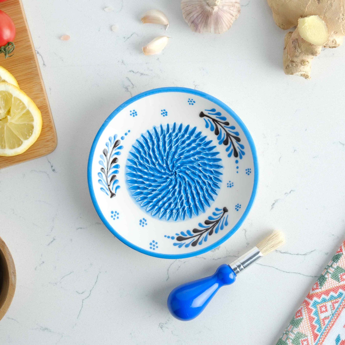 Decorative ceramic garlic grater plate with blue floral design on a white marble surface with kitchen items.