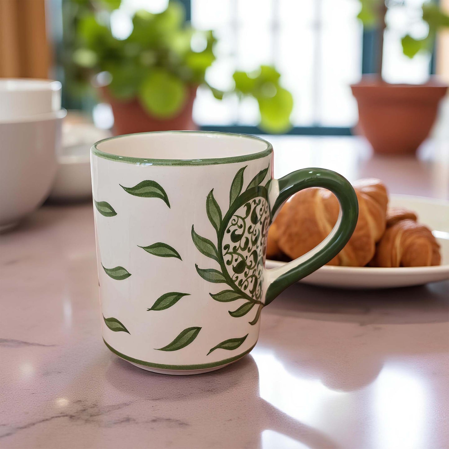White mug with green leaf pattern on a marble surface with pastries and plants in the background