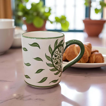 White mug with green leaf pattern on a marble surface with pastries and plants in the background