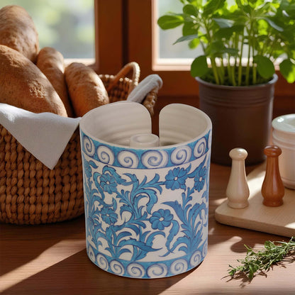 Decorative blue and white ceramic paper towel holder on a wooden surface with plants and bread in the background.