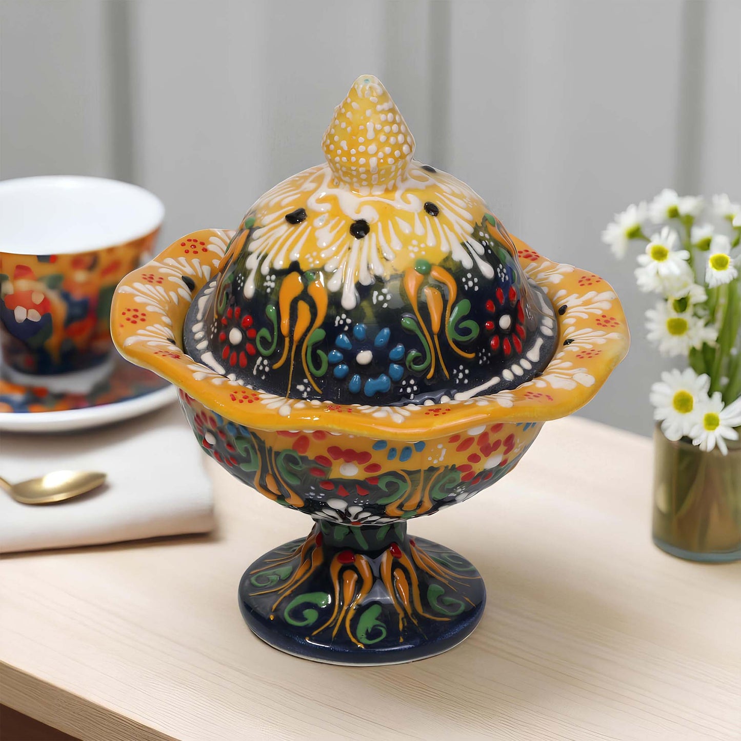 Colorful ceramic sugar bowl with a lid on a table with a cup and flowers in the background