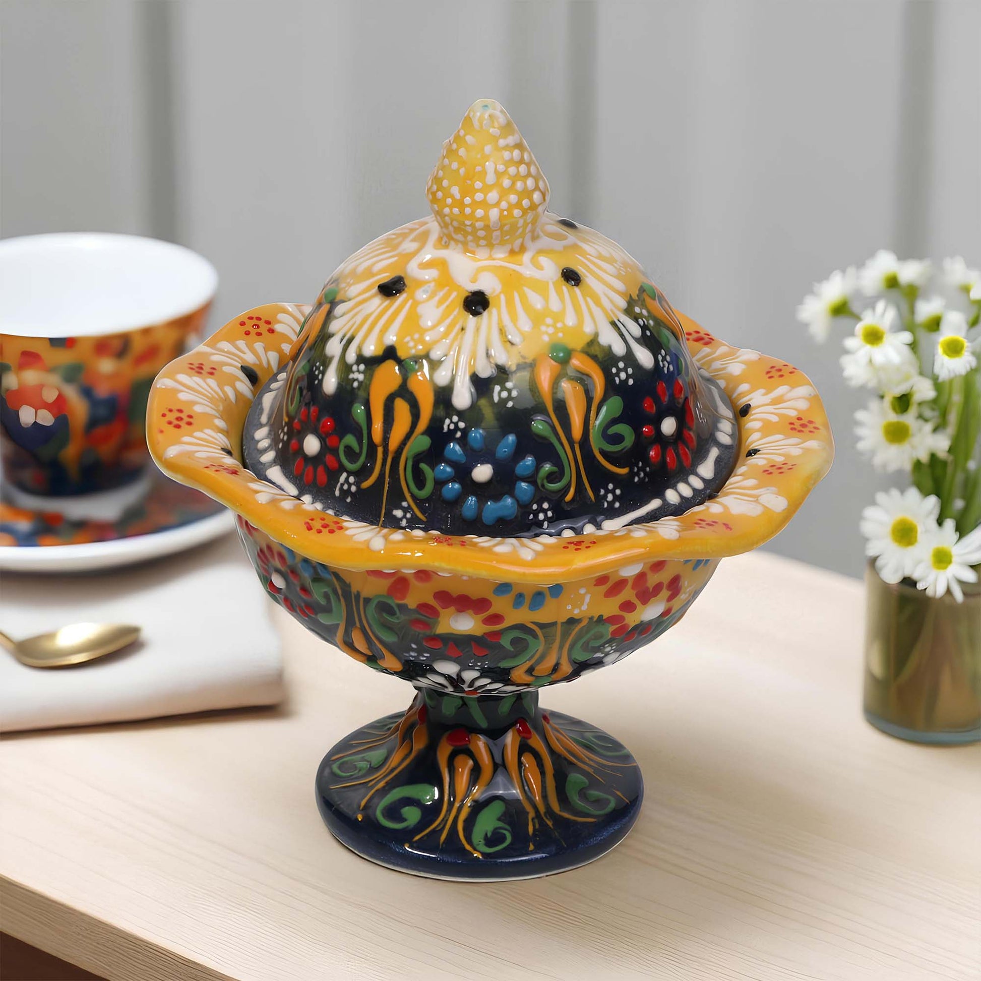 Colorful ceramic sugar bowl with a lid on a table with a cup and flowers in the background