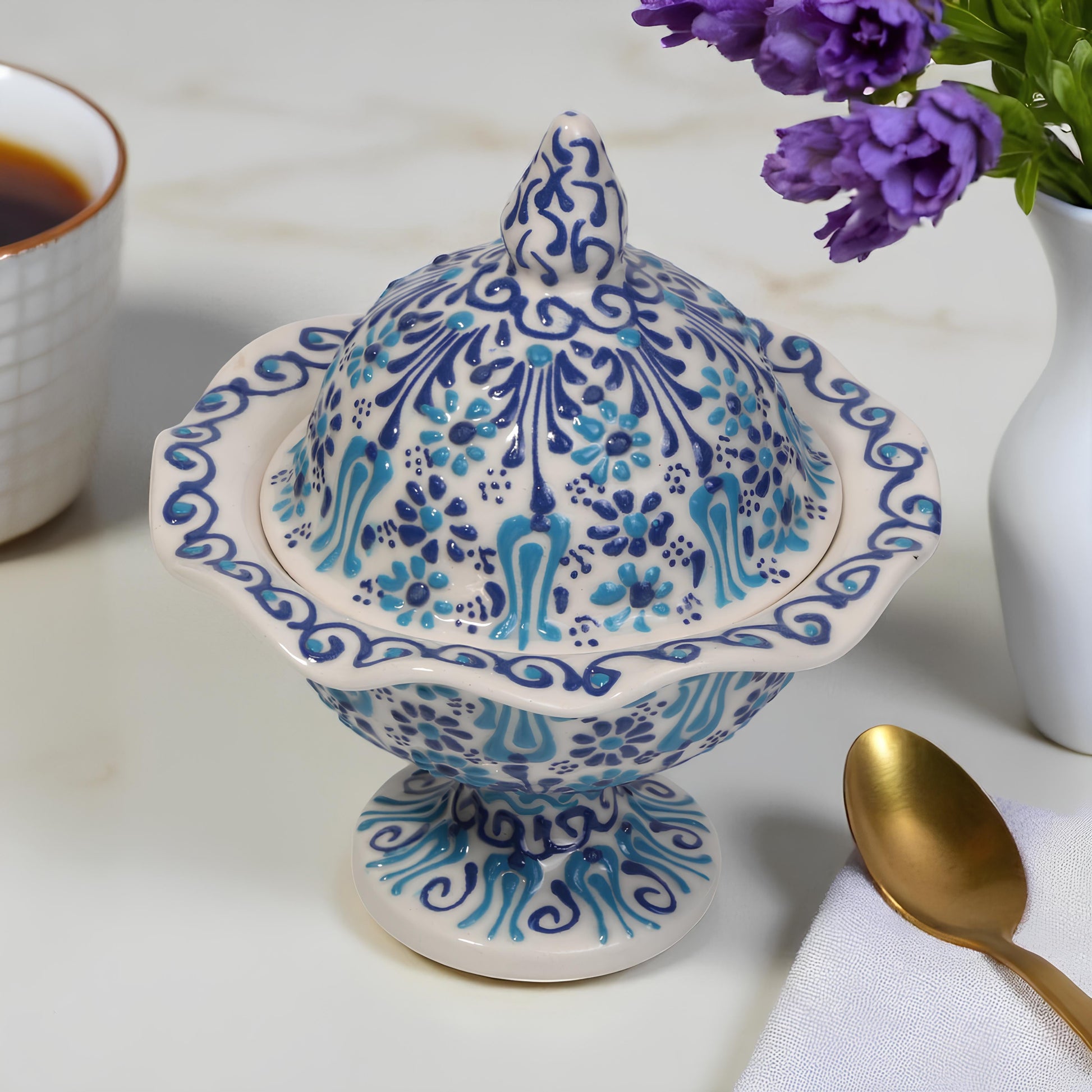 Decorative ceramic sugar bowl with blue floral patterns on a white surface with a cup of coffee and flowers.