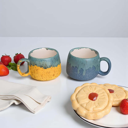 Two ceramic mugs with a yellow and blue gradient on a table with cookies and strawberries.