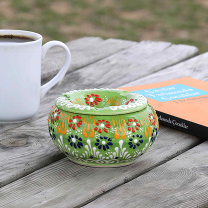 Decorative green ashtray with floral patterns on a wooden surface next to a white mug and book.