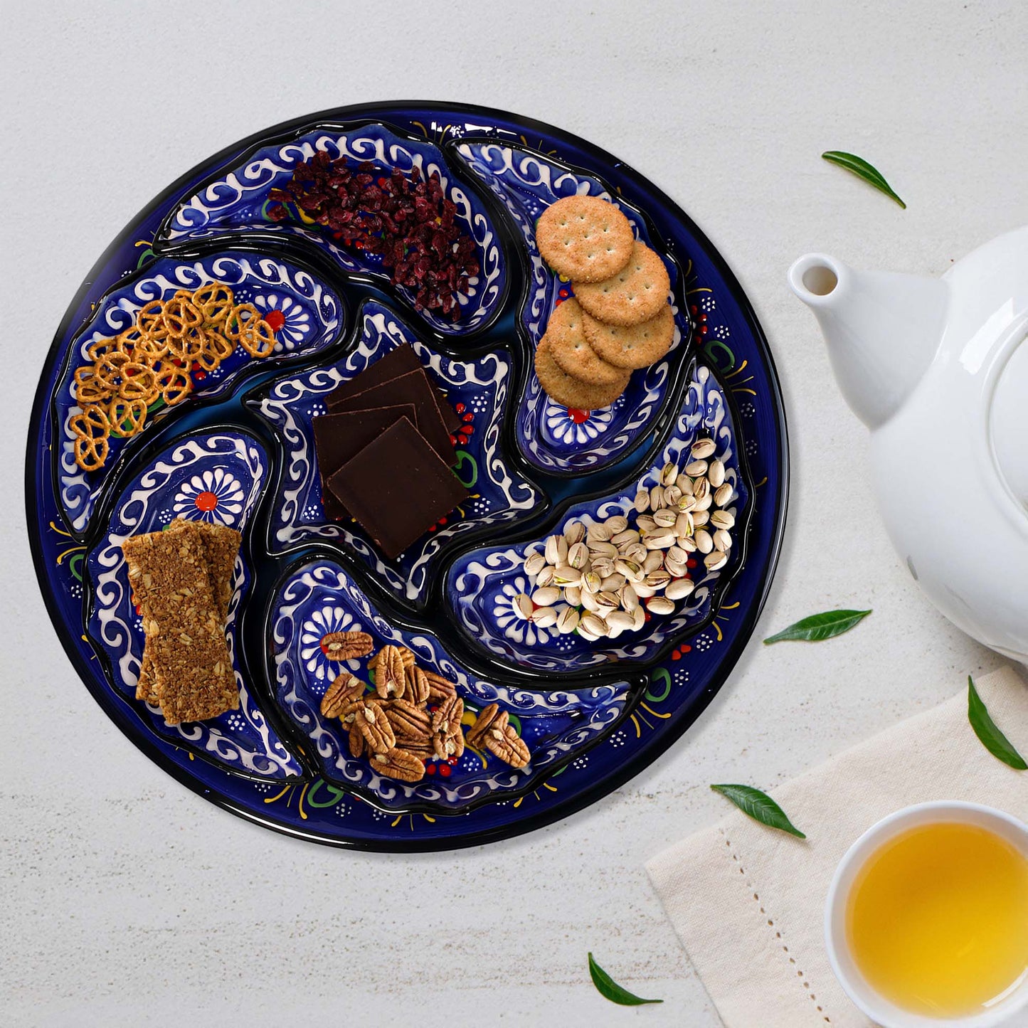 Decorative snack serving tray set with assorted snacks on a white surface with a teapot and cup of tea.