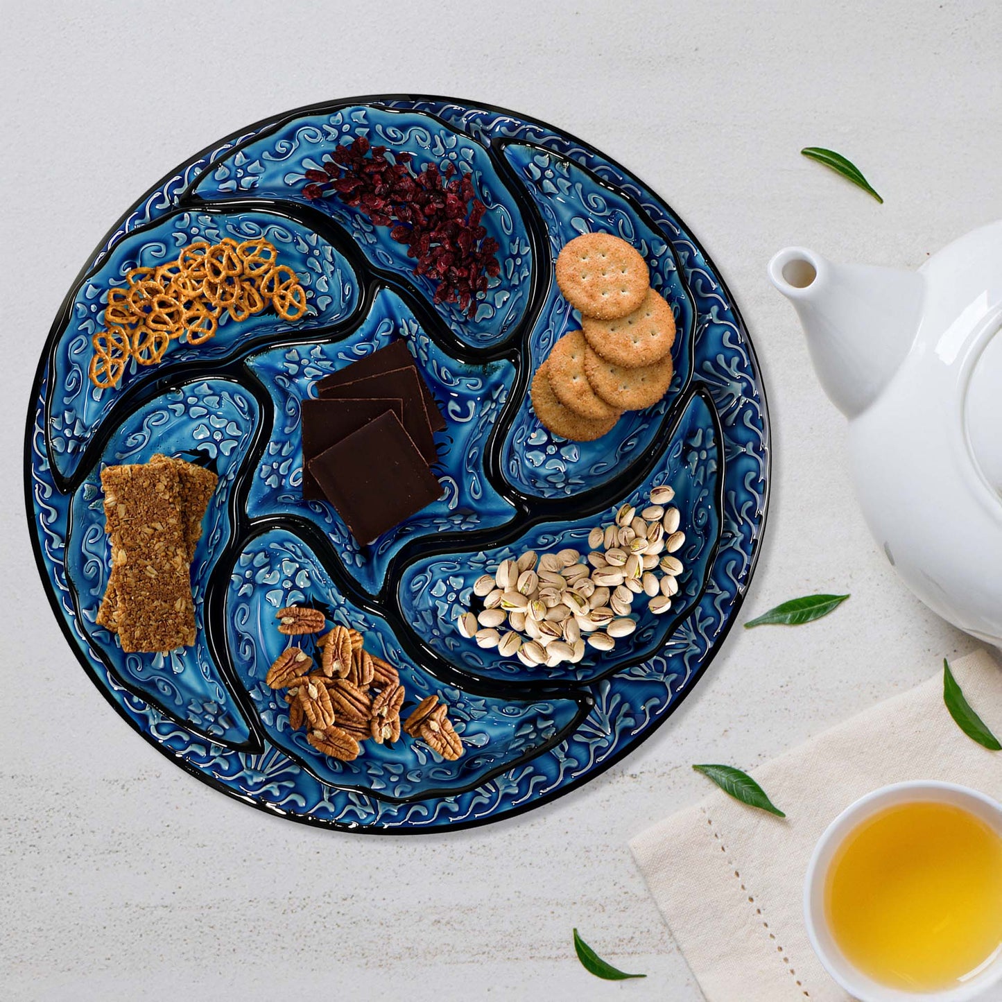 Decorative blue snack serving tray set with snacks on a white surface with a teapot and cup.