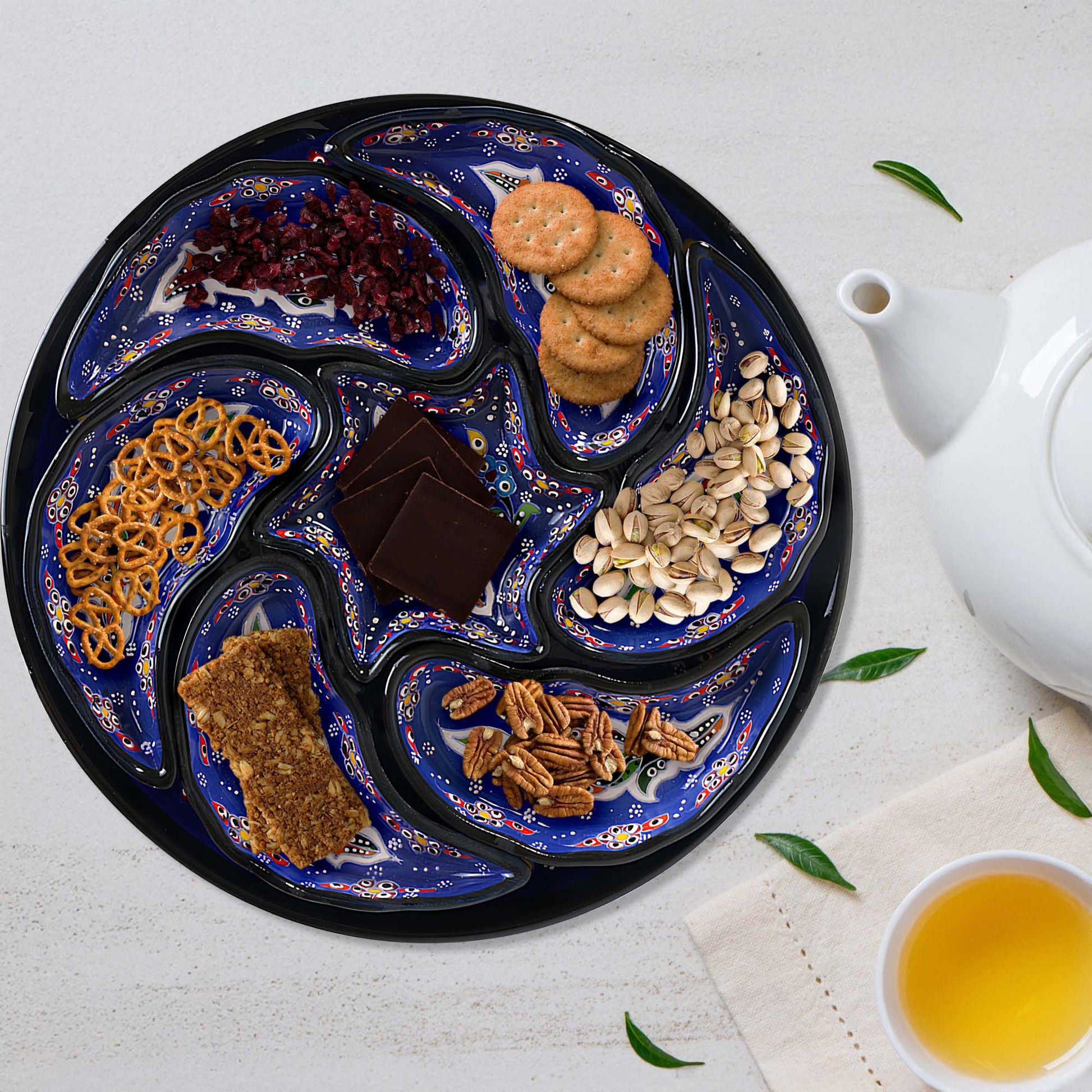 Decorative tray with various snacks on a white surface with a teapot and cup.