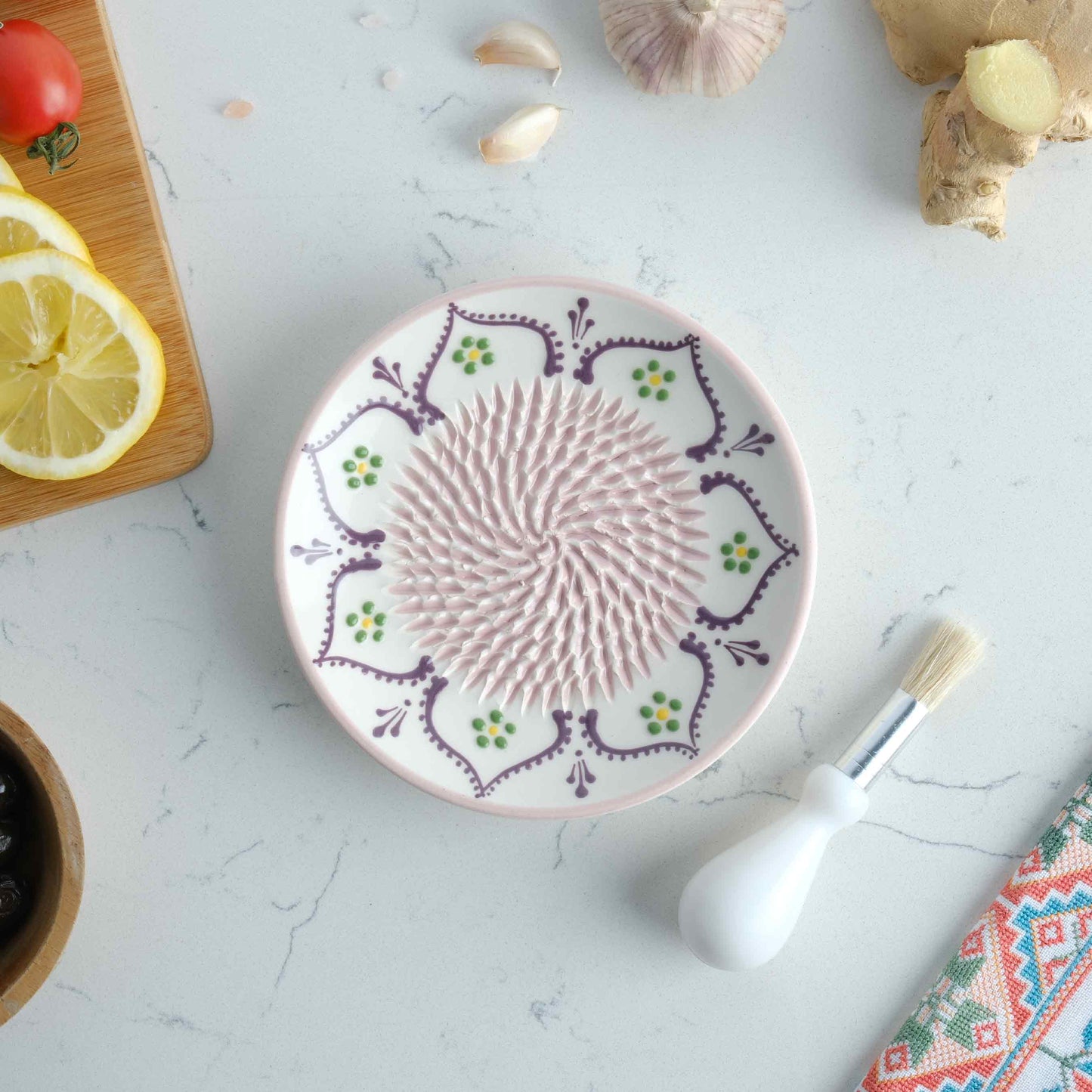 Decorative ceramic garlic grater plate with floral patterns on a kitchen counter with ingredients and tools.