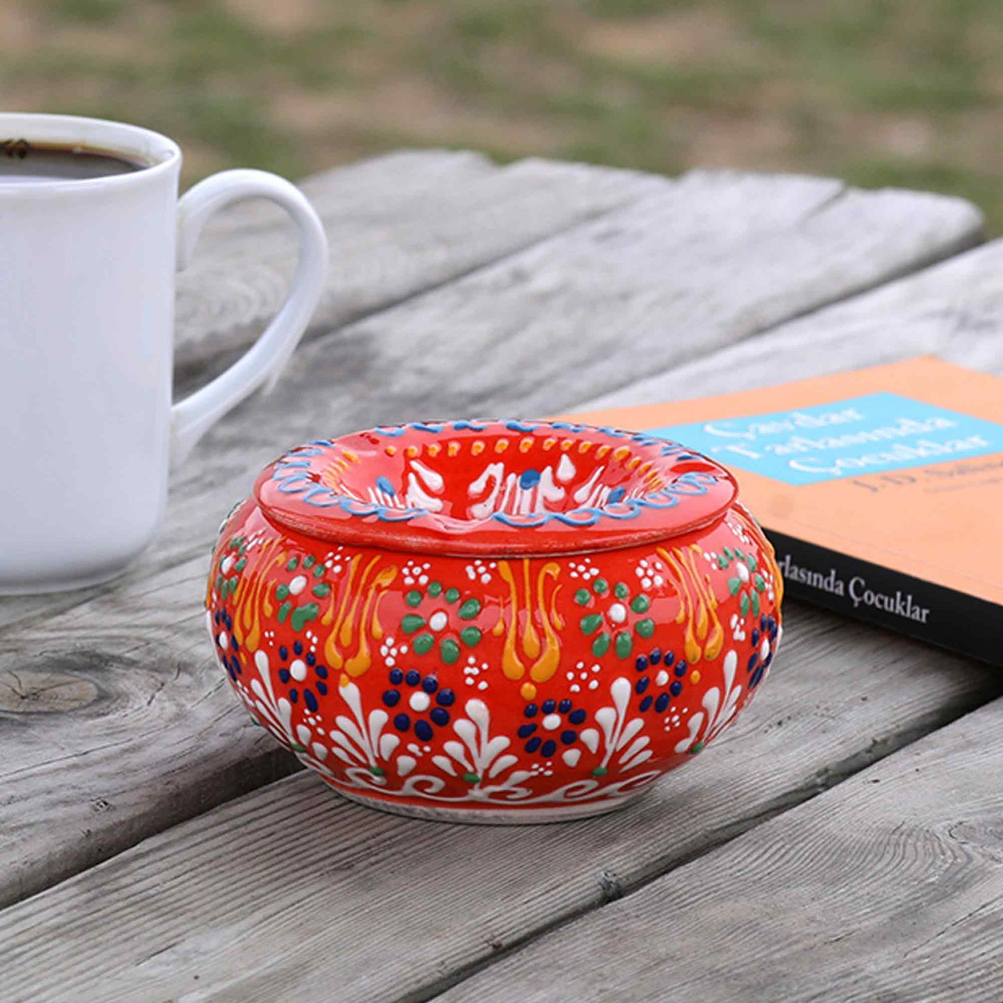 Decorative ceramic ashtray with colorful patterns on a wooden surface next to a white mug and book.