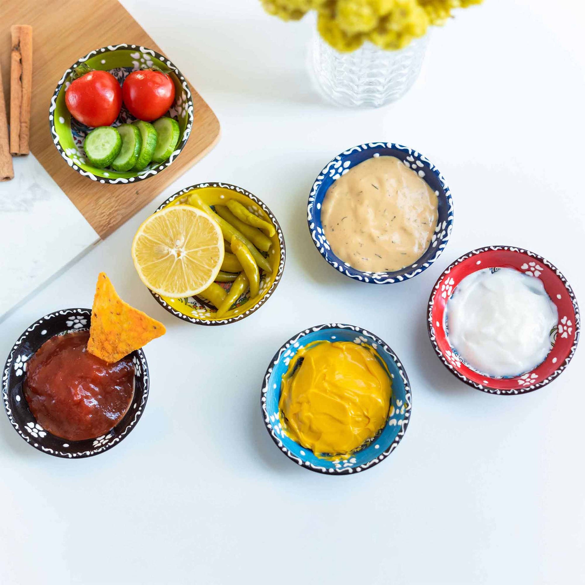 Assorted condiments in small dip bowls on a white surface with a wooden board and flowers in the background.