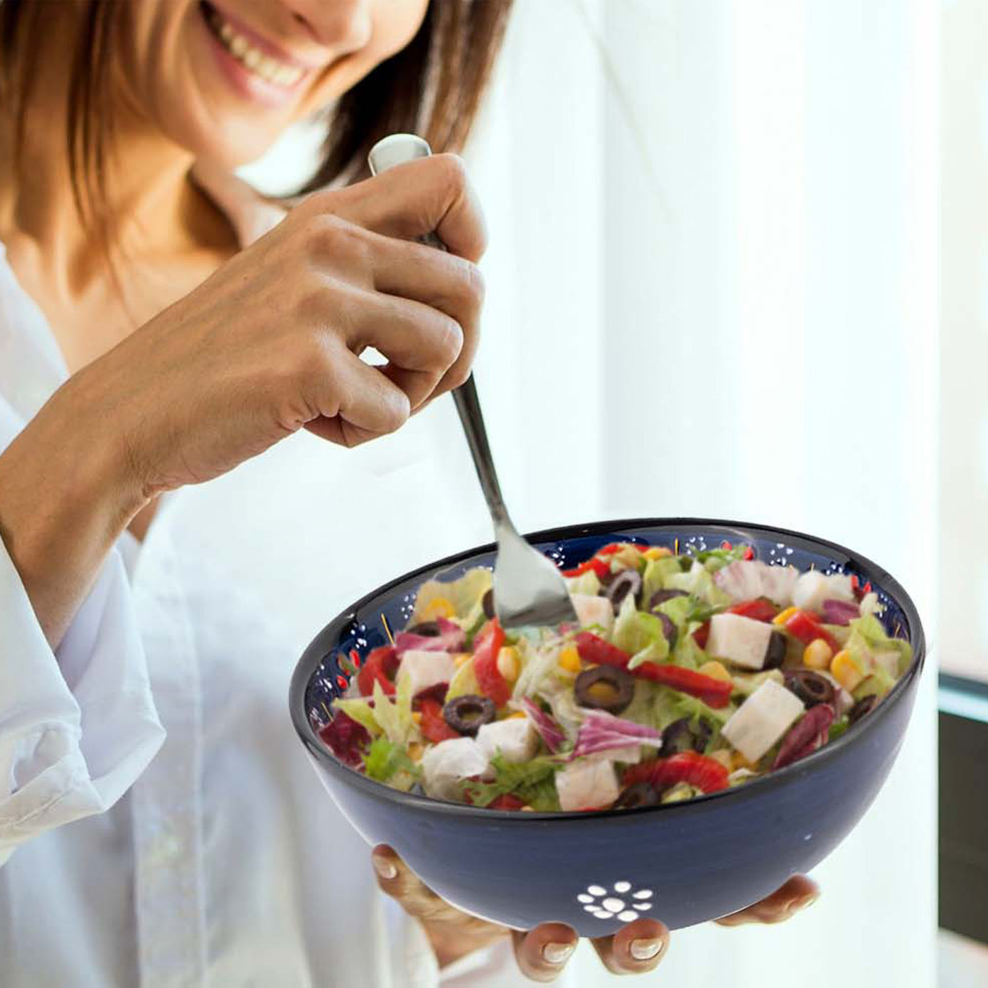 Person holding a blue ceramic bowl of salad with a fork, smiling.
