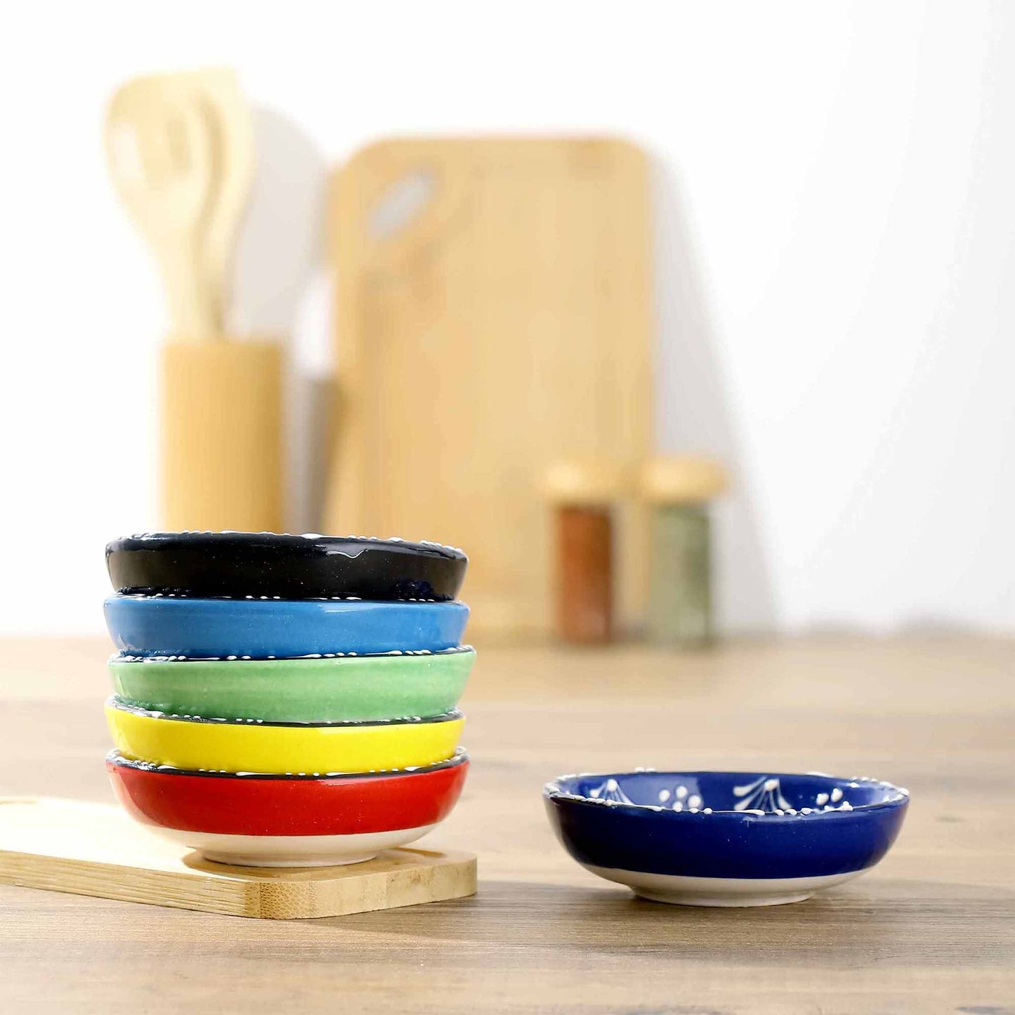 Stack of colorful ceramic dip bowls on a wooden surface with a blurred kitchen background.